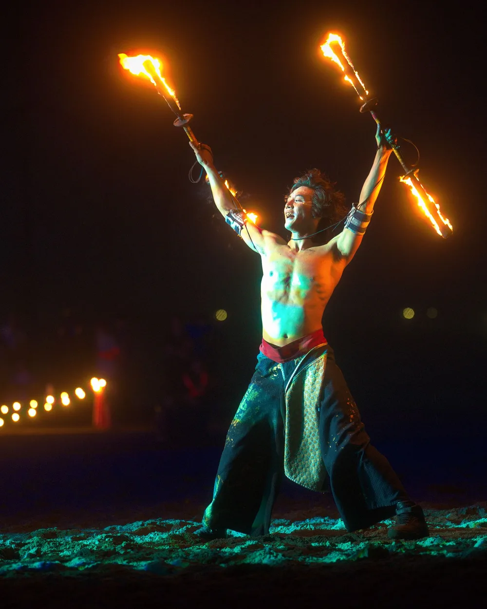 A fire performer on a beach at night, both torches raised, with a crowd behind him — representative photograph, not Ka Moana's own stage
