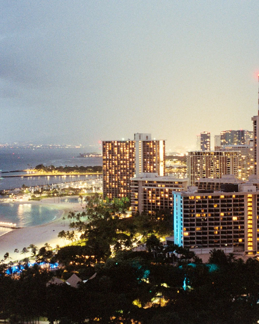 The Honolulu waterfront skyline lit up at dusk, seen across the water from the beach (representative photograph of Honolulu at dusk, not Ka Moana's own lawn)